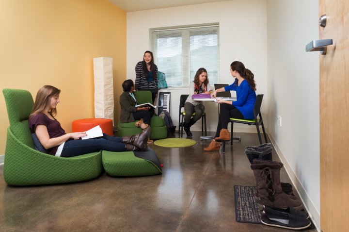 A group of five students gather in a cozy study room. Two are seated on green lounge chairs, one stands by the window, and two others sit at a small table working on papers. Boots and shoes are placed near the door, and the room has a relaxed atmosphere with soft lighting and a yellow accent wall.